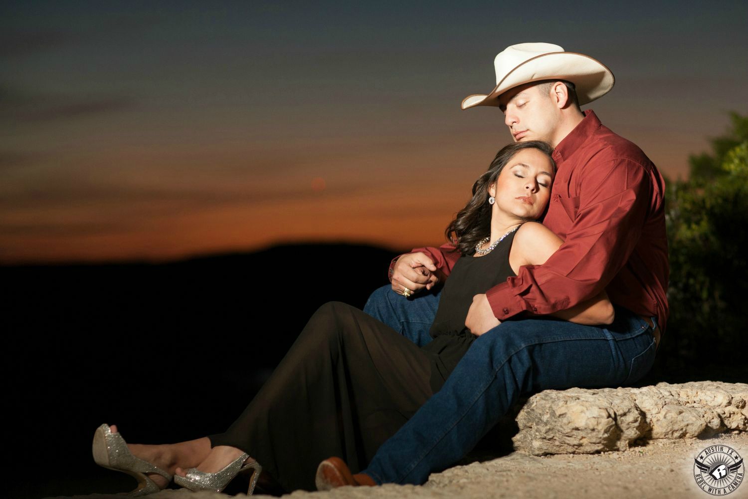 Dark haired Latino girl wearing a long black dress and a jeweled necklace and diamond covered silver heels snuggles up to a guy wearing a tan cowboy hat a and a maroon long sleeve button up shirt and blue jeans sitting on a ledge in front of an orange cloudy sunset at the 360 Bridge   overlook in this soft engagement portrait in Austin.